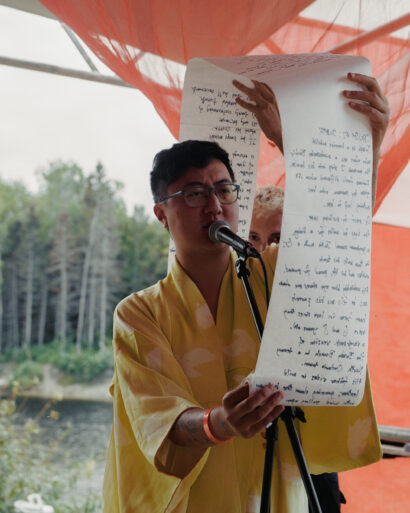 A person stands outdoors, speaking into a microphone while holding a long scroll of handwritten text that curls at the top. They wear glasses and a yellow robe with light-colored patterns. Behind them, red fabric drapes create a vivid backdrop, and trees and a body of water are visible in the background.