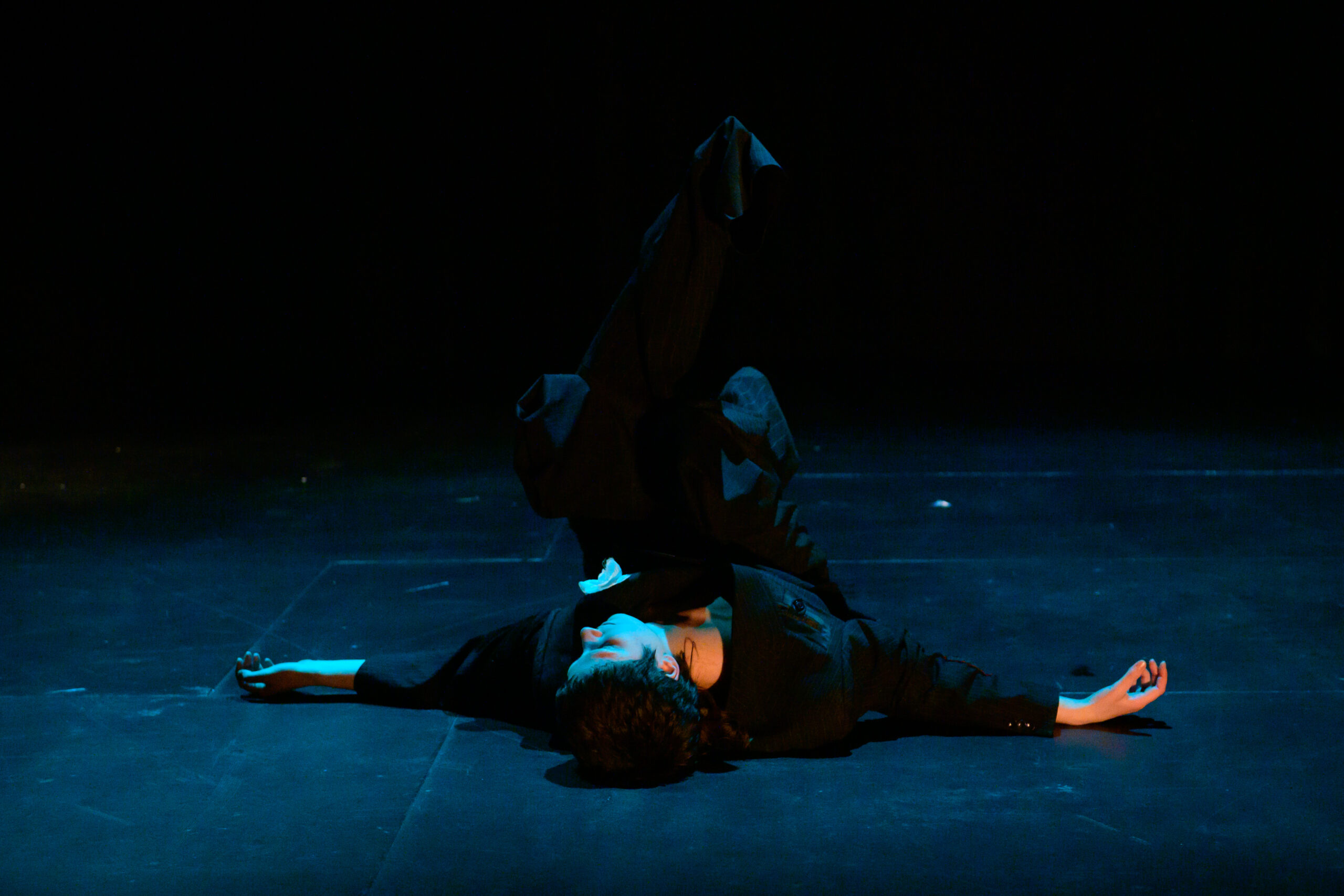 Image of performer in an black oversized suit, lying on their back with their legs in the air.