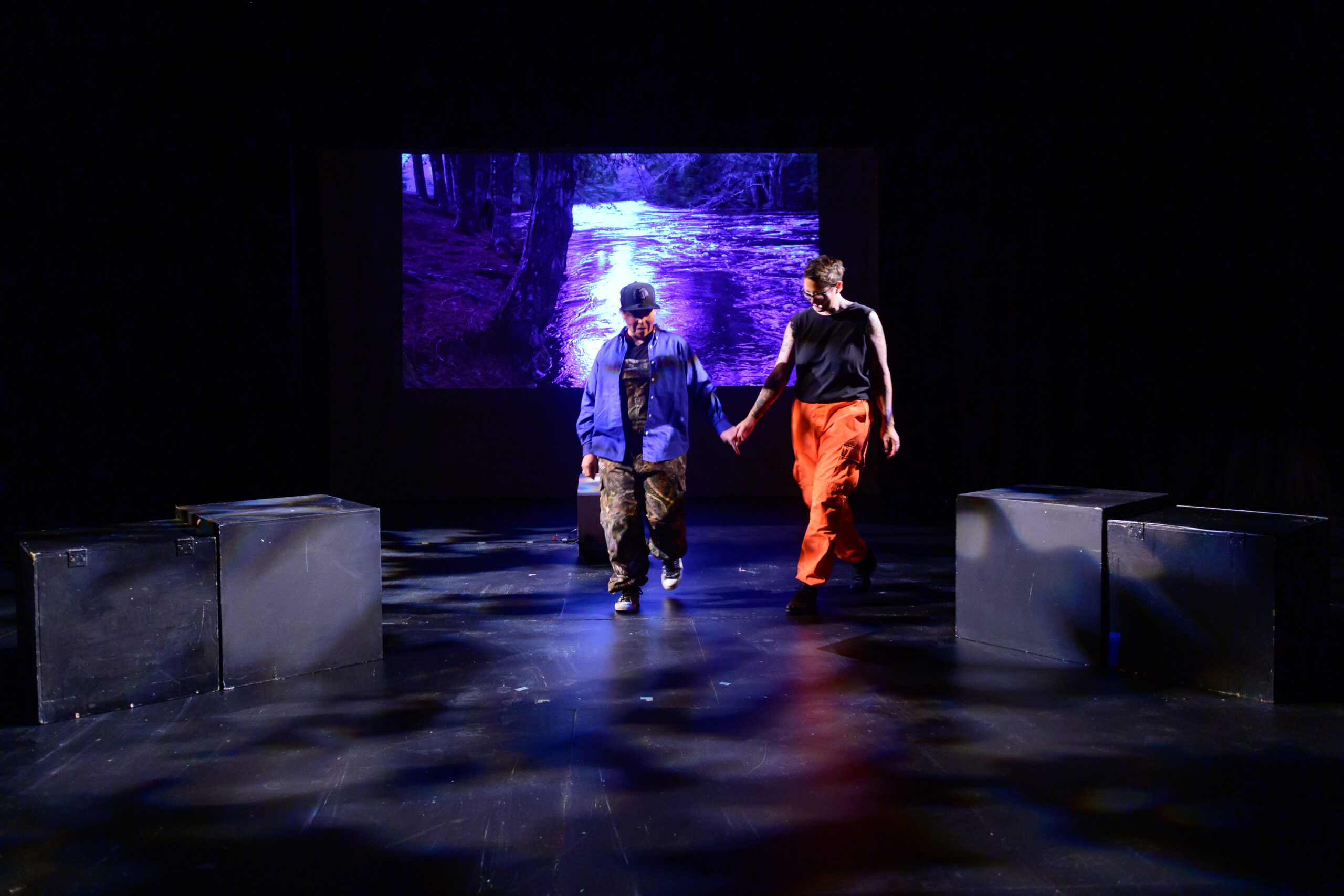 Image of two performers on stage walking and holding hands. Large black cubes are on either side of the stage and a river is projected behind them.