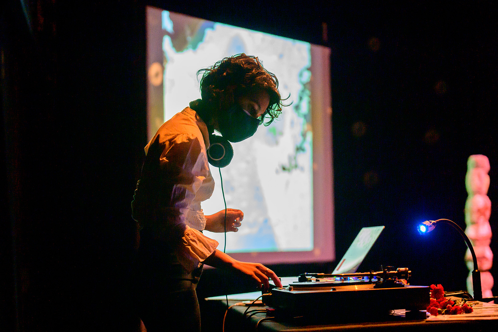 A person with short hair stands in front of a laptop and DJ setup. They are wearing a white blouse, headphones, and a face mask. In the background a screen with a projection on it is out of focus.