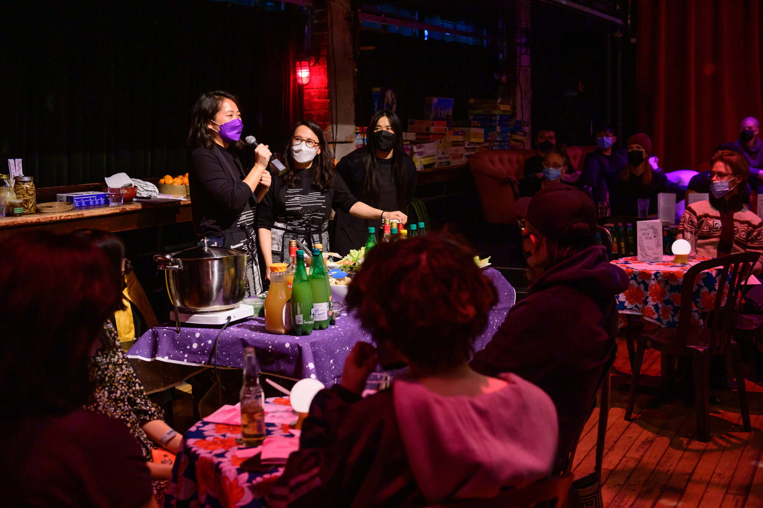 Image of three mashed artists addressing the audience from behind their serving table. The Cabaret is full of audience members seated with drinks and food at tables or on couches.
