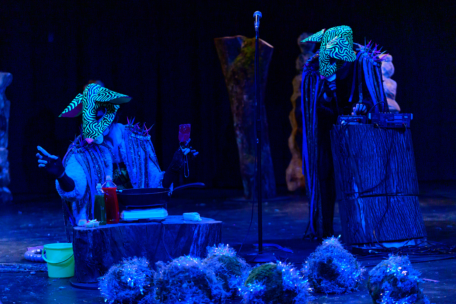 Two figures wearing neon rat masks in a theatre space. The one on the left kneels in front of a tree stump - atop it are a hot plate and frying pan and two bottles of soap. The figure on the right crouches over a tree stump that holds a small sound board and a microphone.