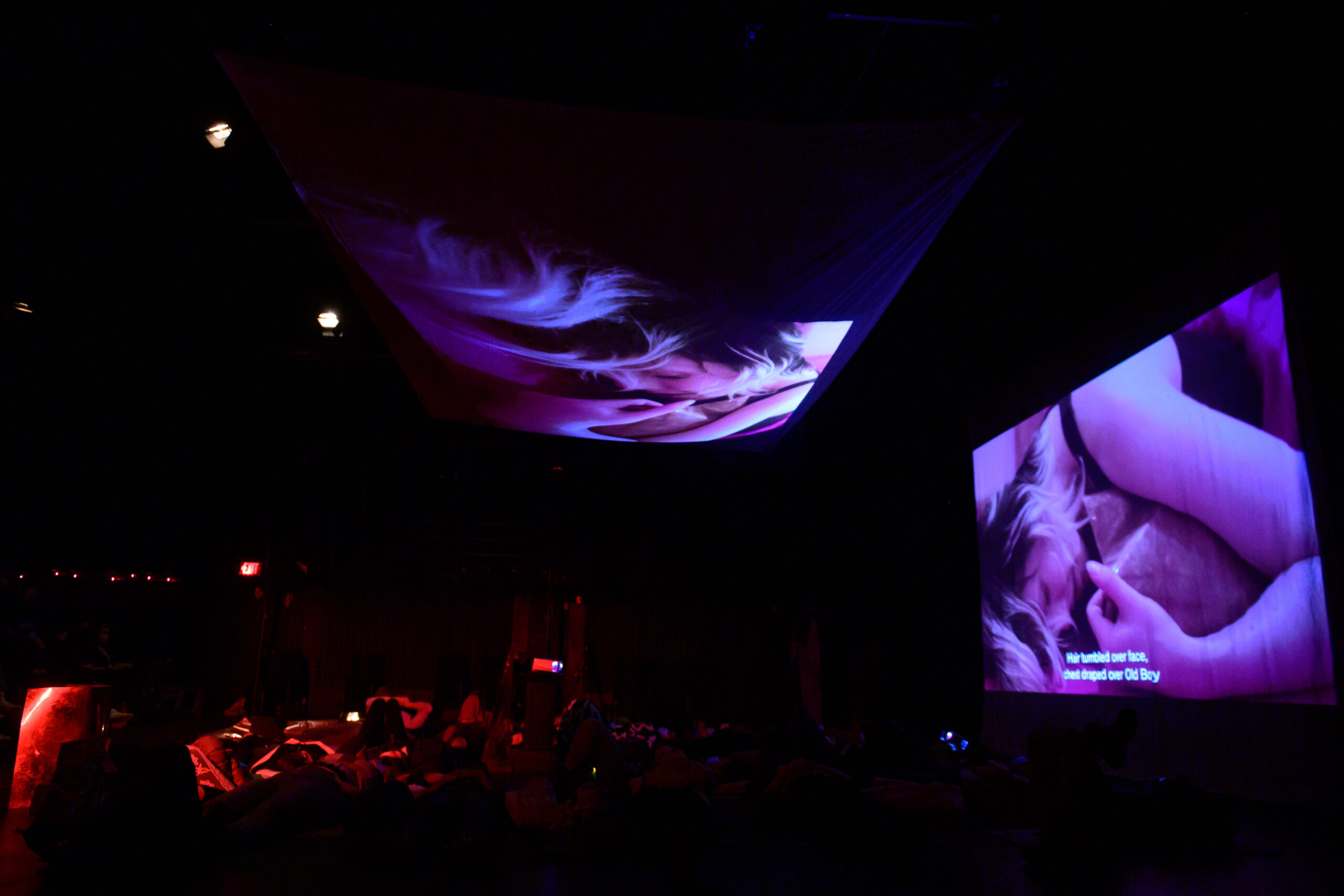 Image of two large projections of performer Laura Fisher cradling their art. Audience members lie on mats and pillows on stage.