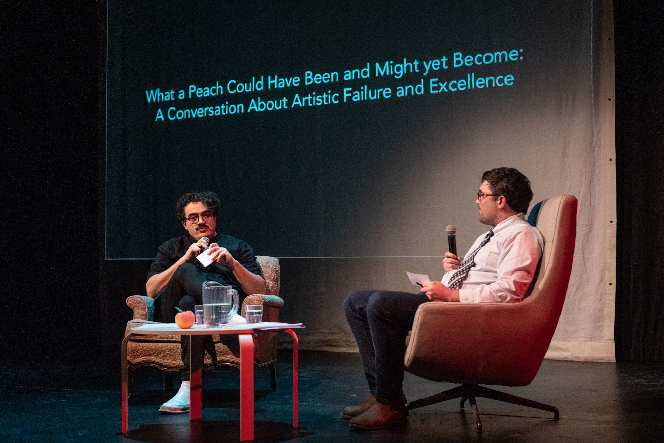 Image of Julian and Henry sitting in armchairs on stage with a table in between them. There is a jug of water, two water glasses, and a peach on the table. Both Julian and Henry speak into microphones and hold cue cards. Projected on a screen behind them is the title of their project; “What a Peach Could Have Been and Might yet Become: A Conversation About Artistic Failure and Excellence”