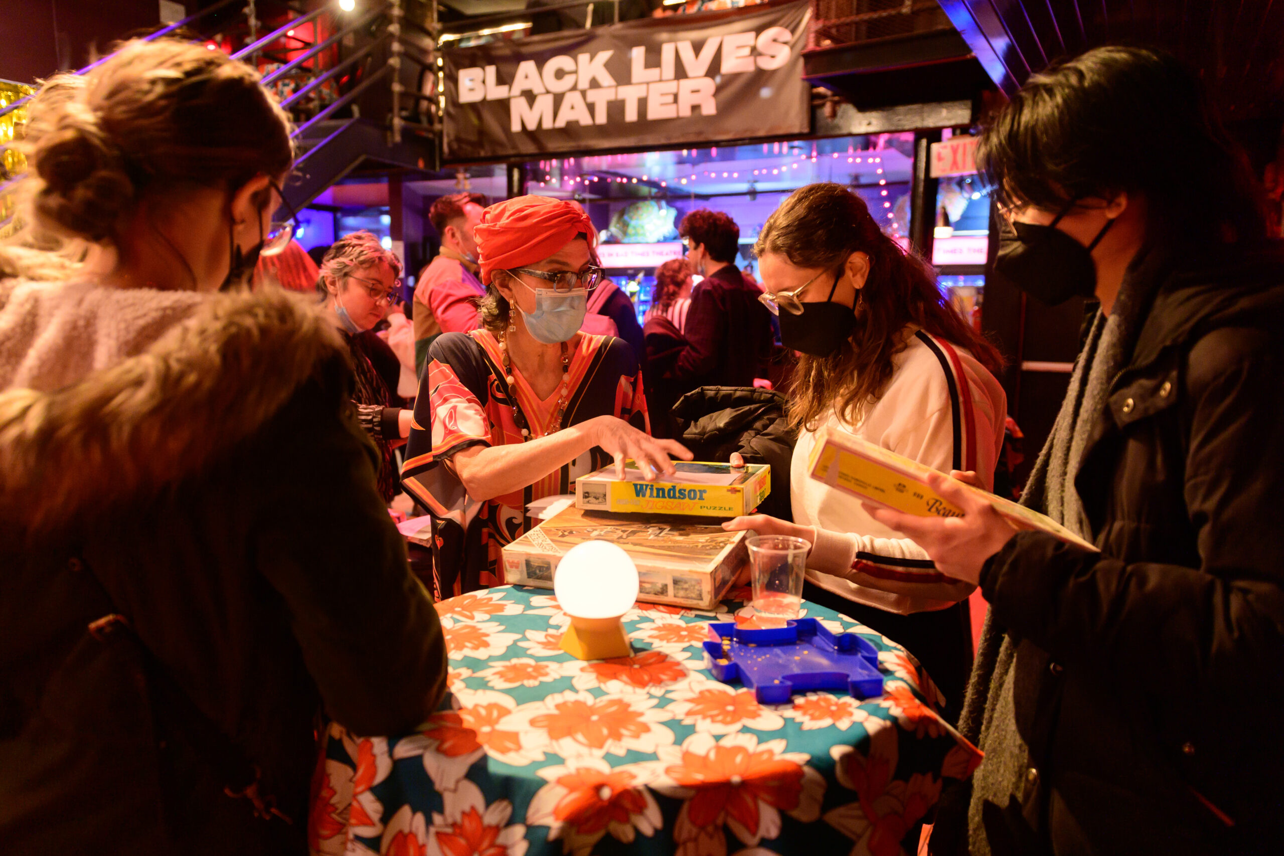 Image of a masked artist holding a puzzle and engaging with audience members in the Cabaret’s Puzzle Lounge.