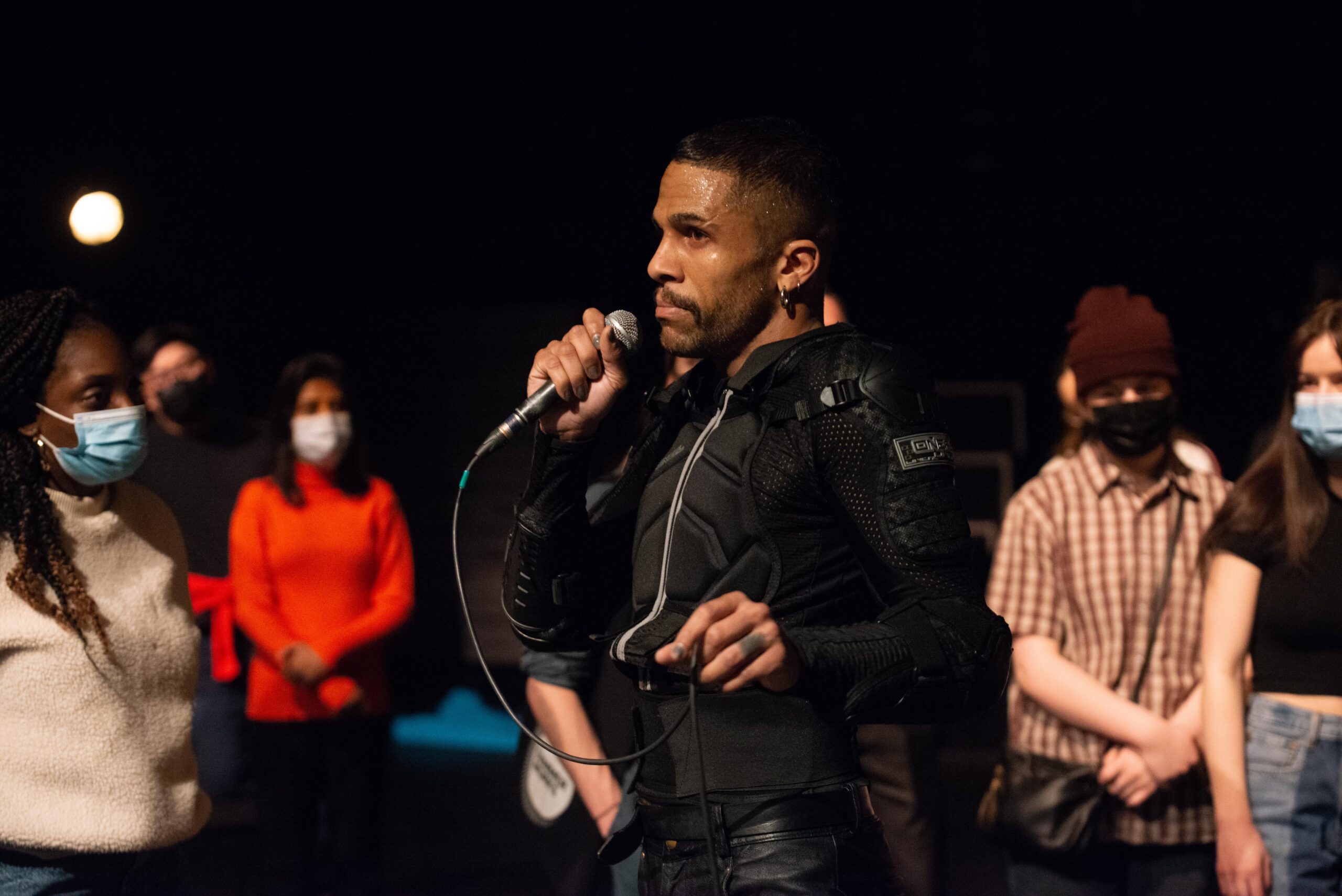 Image of performer holding a microphone, walking among a masked audience, wearing a black jacket.