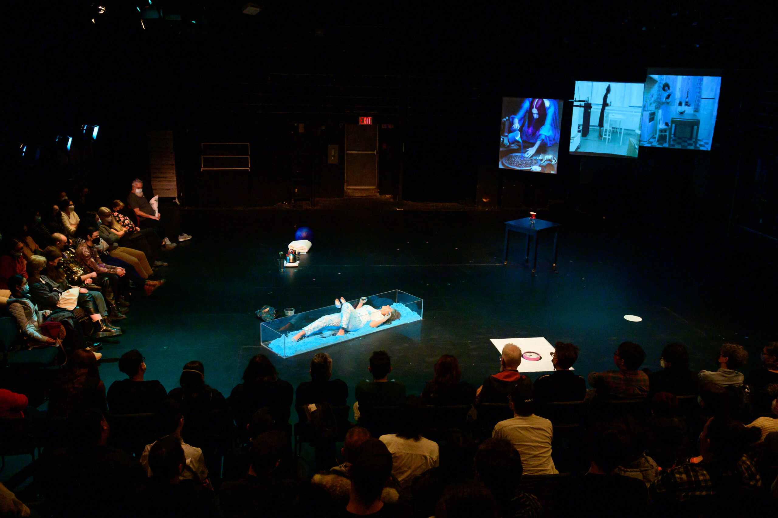 Image of performer in a glass rectangle container on stage. The performer is wearing all white, and is lying in a bright blue powder. A kettle, blanket, and ball sit on the stage floor behind the performer, as well as a small black table with a coffee mug and filter. In the background three large screens depict images of kitchen/housework. 