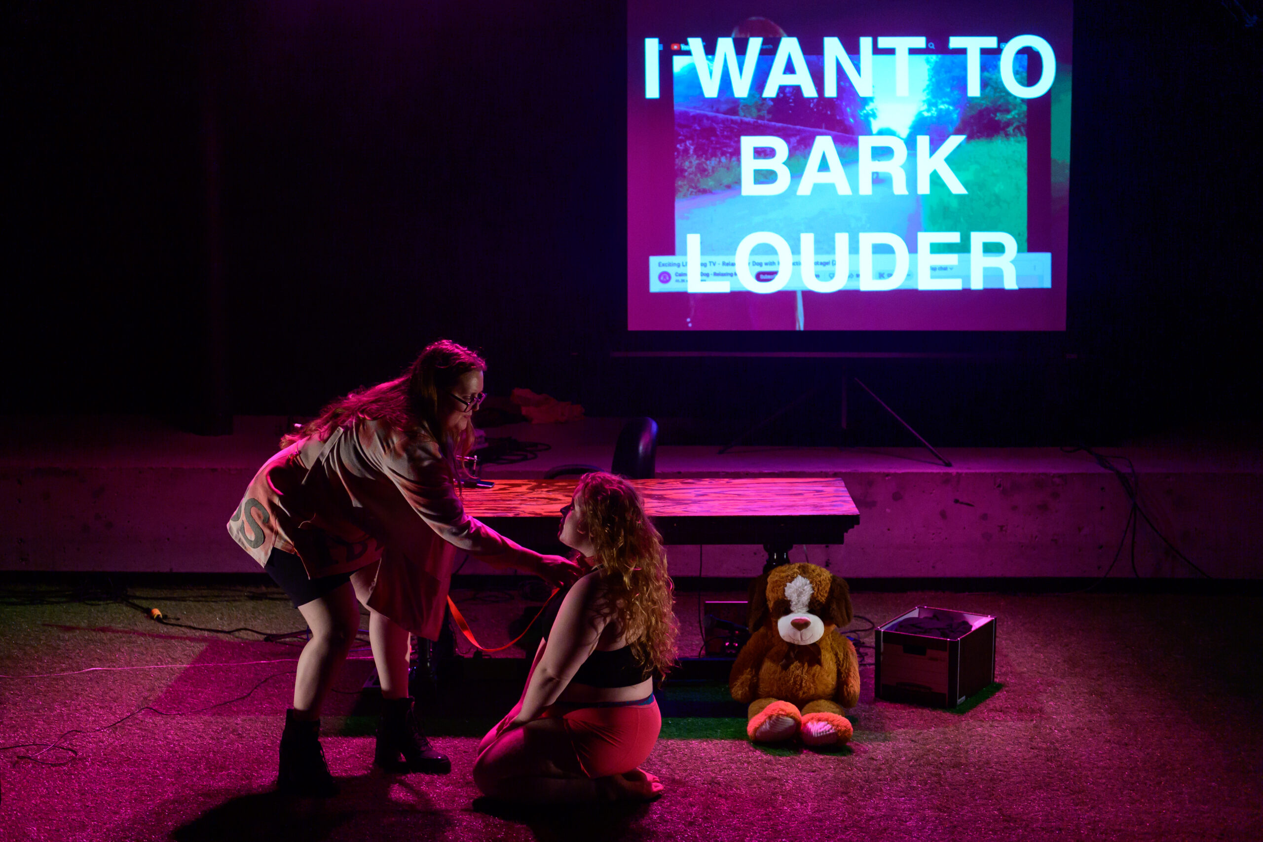 Image of Dasha and Gislina on stage; a projection with large texts reads, I WANT TO BARK LOUDER in the background, Dasha stands over Gislina, who is kneeling, and holds them by his collar. A stuffed dog and a box of files sit on the floor next to a table behind them.