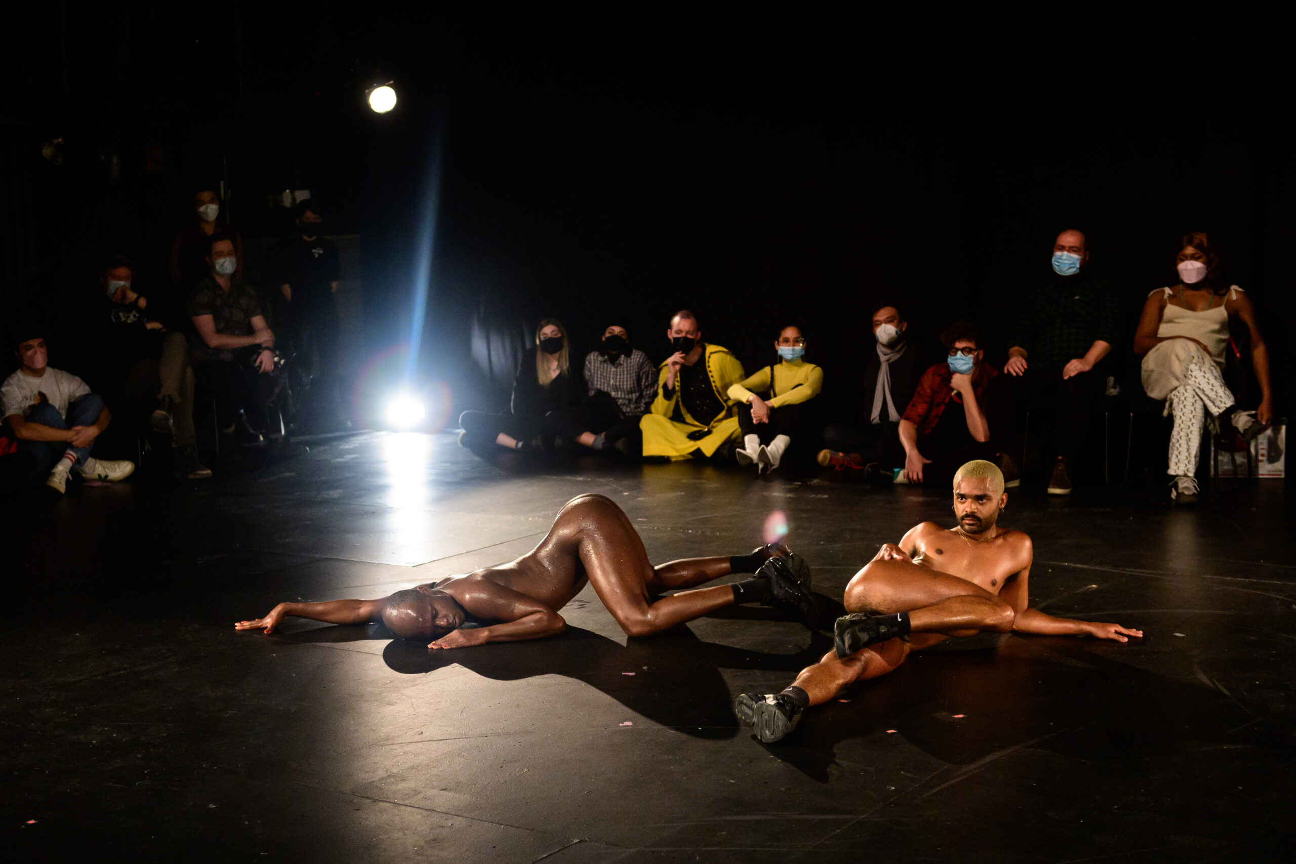 Image of performers Davi and Wallace posing on the stage floor. They are wearing black shoes and socks. Audience sits behind them in chairs and on the floor, masked. A bright light sits on the floor in the top left corner of the stage.