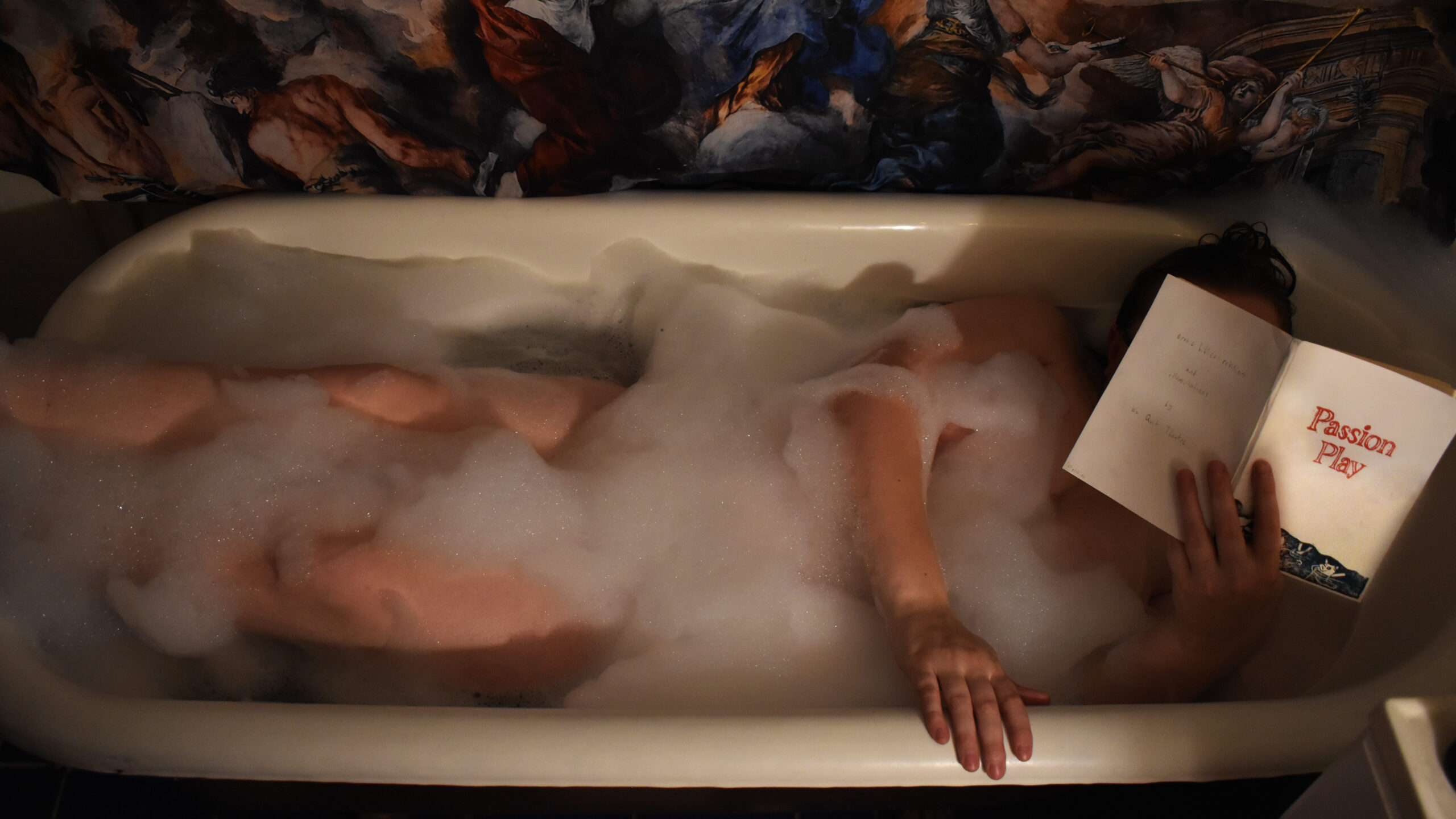 Image of Dasha lying in a bubble bath. She is holding a book open in front of her face titled, "Passion Play"
