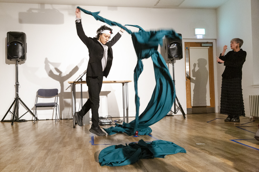 Image of performer Claudia Edwards. Claudia is lifting a long, teal piece of fabric, wearing a black jacket and pants, and a white headband, in a studio space. In the background are two standing speakers, a chair, table, and an ASL interpreter, wearing all black.
