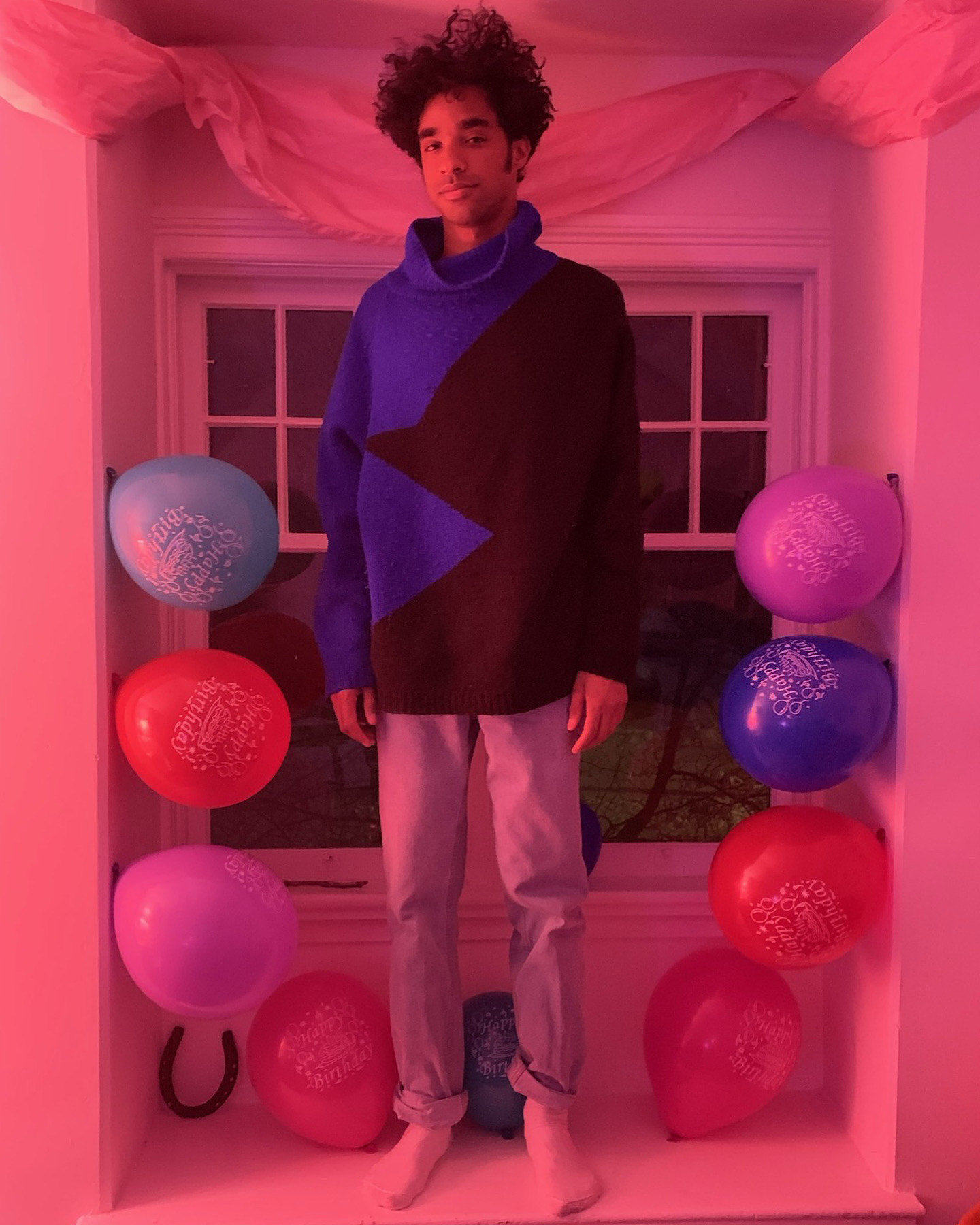 Stephen stand in a window sill with their arms at their sides. They wear a blue and black colour-blocked sweater, and jeans rolled at the cuffs. Behind and around them are streamers and balloon in different colours that say "Happy Birthday"