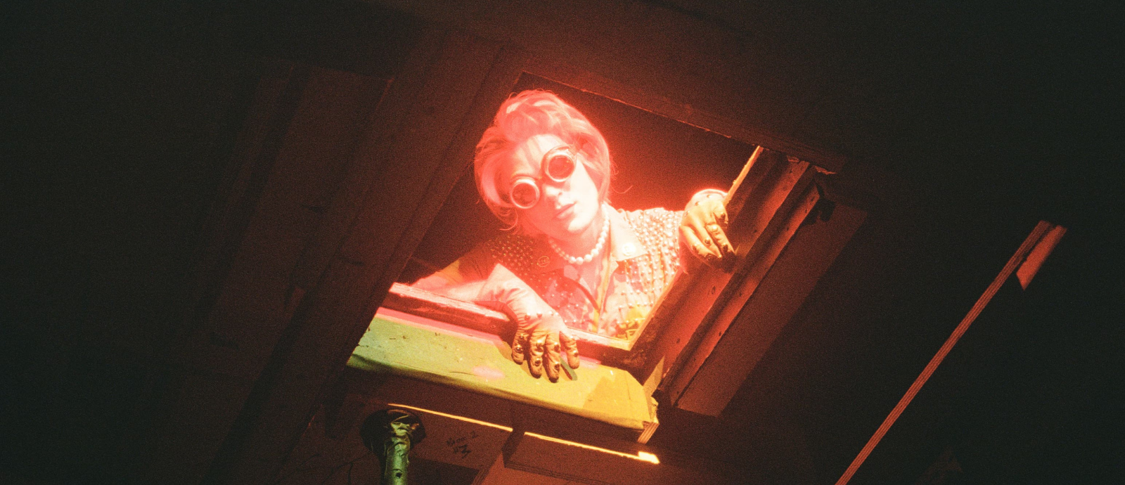 Pearle looks down at the camera through a trapdoor. She is wearing stoned leather gloves, a pearl necklace, and goggles.
