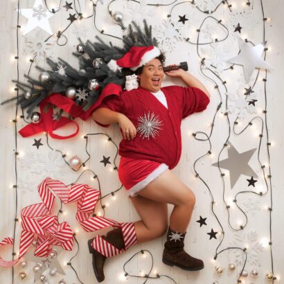 Chris, a tall human with tan skin and black short hair, dons a sexy santa suit. Red velvet shorts paired with a red velvet robe. They hold a small Christmas tree over their head and are laying on a white floor. Around them are white Christmas lights and red rows.