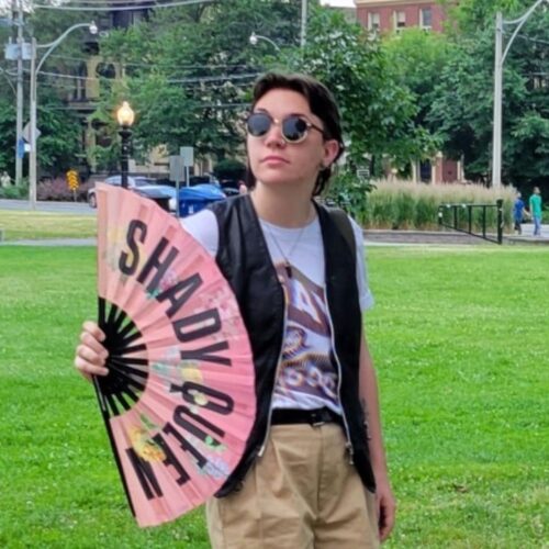Al is standing in a park, with grass and trees behind them. They wear sunglasses, a t-shirt, and a vest, with a fan unfolded in front of them. The fan reads "Shady Queen"