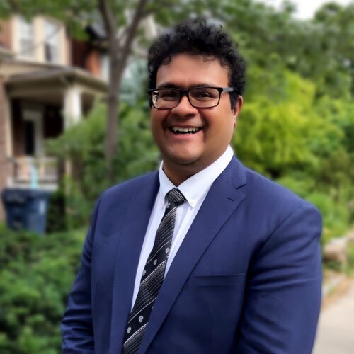 Image of Arjun wearing a navy blue suit with a striped and polka dotted black and gold tie. He is wearing glasses. Trees and houses are out of focus in the background.