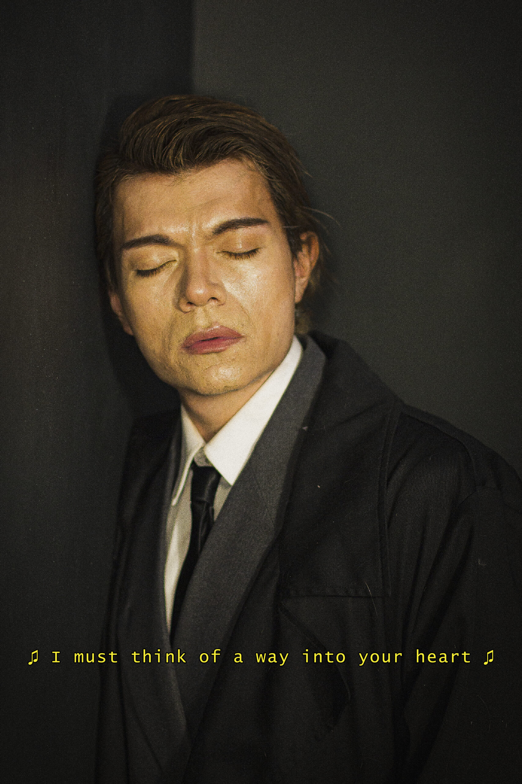 Jaime wears a white shirt, black tie, grey blazer, and black overcoat. He leans against a wall with his eyes closed. At the bottom of the image text bracketed by music notes reads "I must think of a way into your heart"