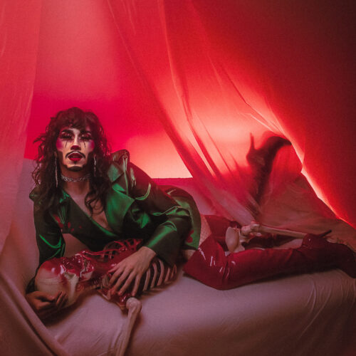 Image of Heath lying on their front, embracing a skeleton. They are wearing drag makeup and an emerald green jacket and tall red heeled boots.
