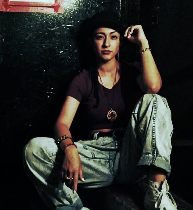 Photo of C4LDEIR4, sitting and posing, wearing light wash jeans and a dark v neck t-shirt.