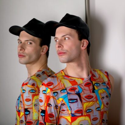 An image of Calvin Cox with his back to a mirror, so that we see him twice. He wears a black ball cap and a colourful shirt with illustrations of faces on it.