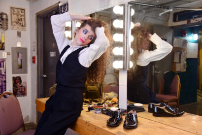 Image of James sitting on the edge of the dressing room table at Buddies. James is wearing a black vest over a white blouse with a ruffled neckline, high waisted black dress pants, and silver heeled boots. They have long, curly brown hair, blue eye shadow, and pink lipstick. Shiny black dress shoes sit on the table next to them, along with all of their stage makeup.