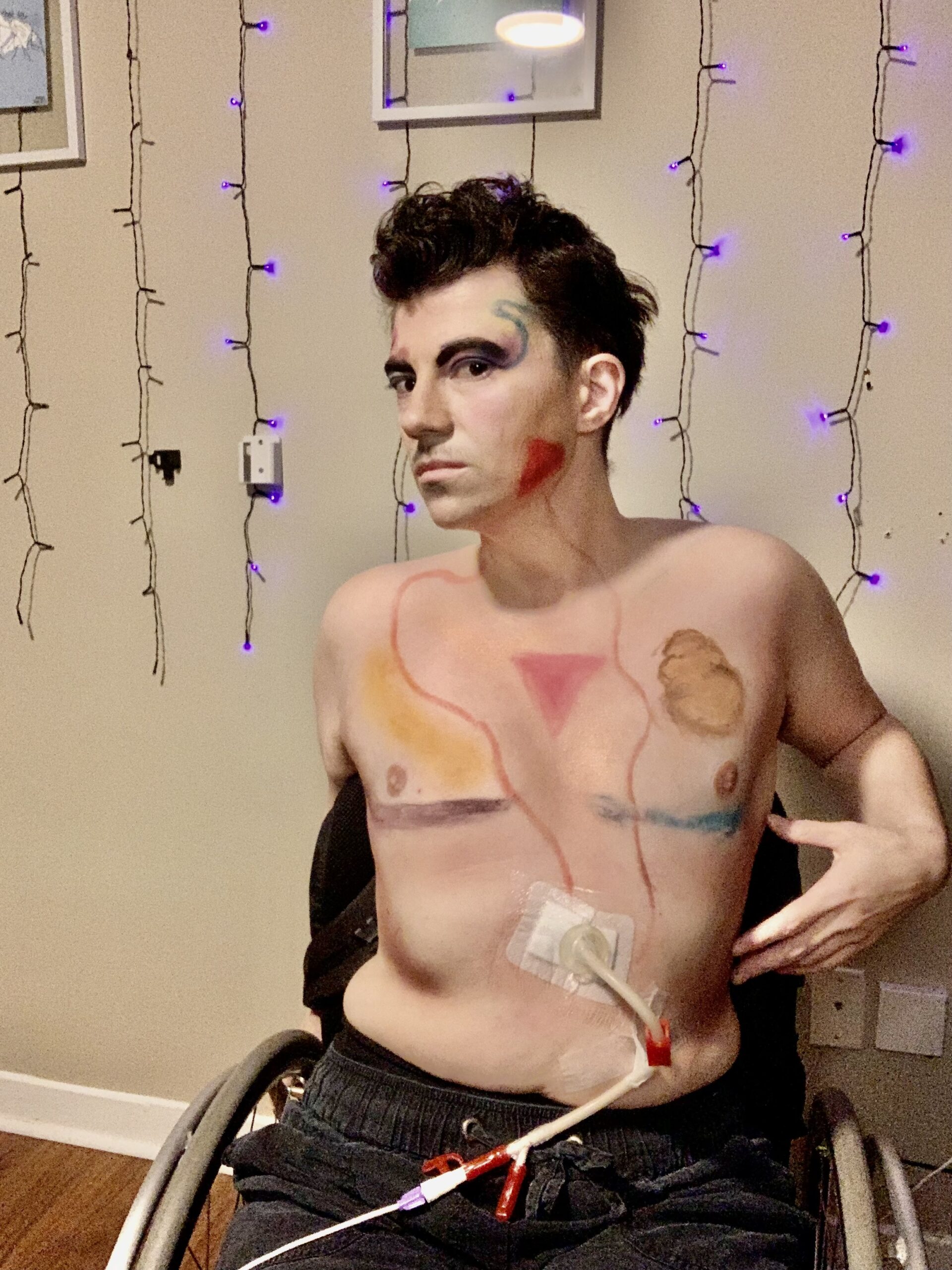 Gabe, Entro-pee, is a white trans masculine person with short brown curly hair using a manual wheelchair. Their top surgery scars visible are highlighted with purple and blue lines. They have a large gastric tube coming from their midline that is connected to smaller line behind them in a backpack for feeding them. On their face , the makeup highlights a small moustache and red triangles on both cheeks with golden highlighting. Their eyes have purple and blue eyeliners in each eye with purple and blue eyeshadows on the eyes with the other colour. The eyeliner twists upwards into tube like shapes to represents the tubes inside and outside of their body. The red triangles on their face is traces downward in swirling lines towards the gastric tubes that feed them. They have a inverted pink triangle in the middle of their chest to represent the queer individuals who the community has lost and will continue to lose to violence. They body is shaded in different shades of golden and white glittering shadows and highlights. They have black tight pants just underneath their midline.