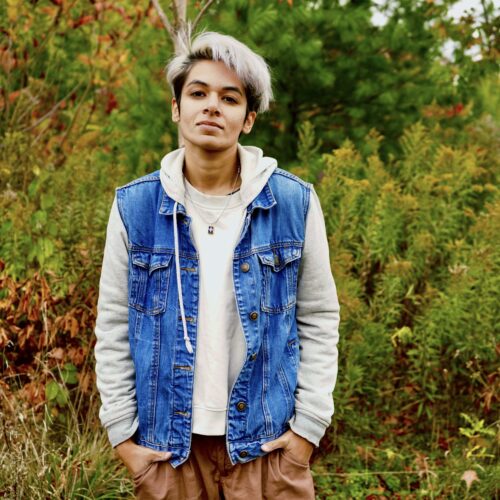 Erum stands in front some shrubs and trees. She has short white hair, with dark roots, and wears a denim vest over a grey hoodie, with her hands in her pockets.