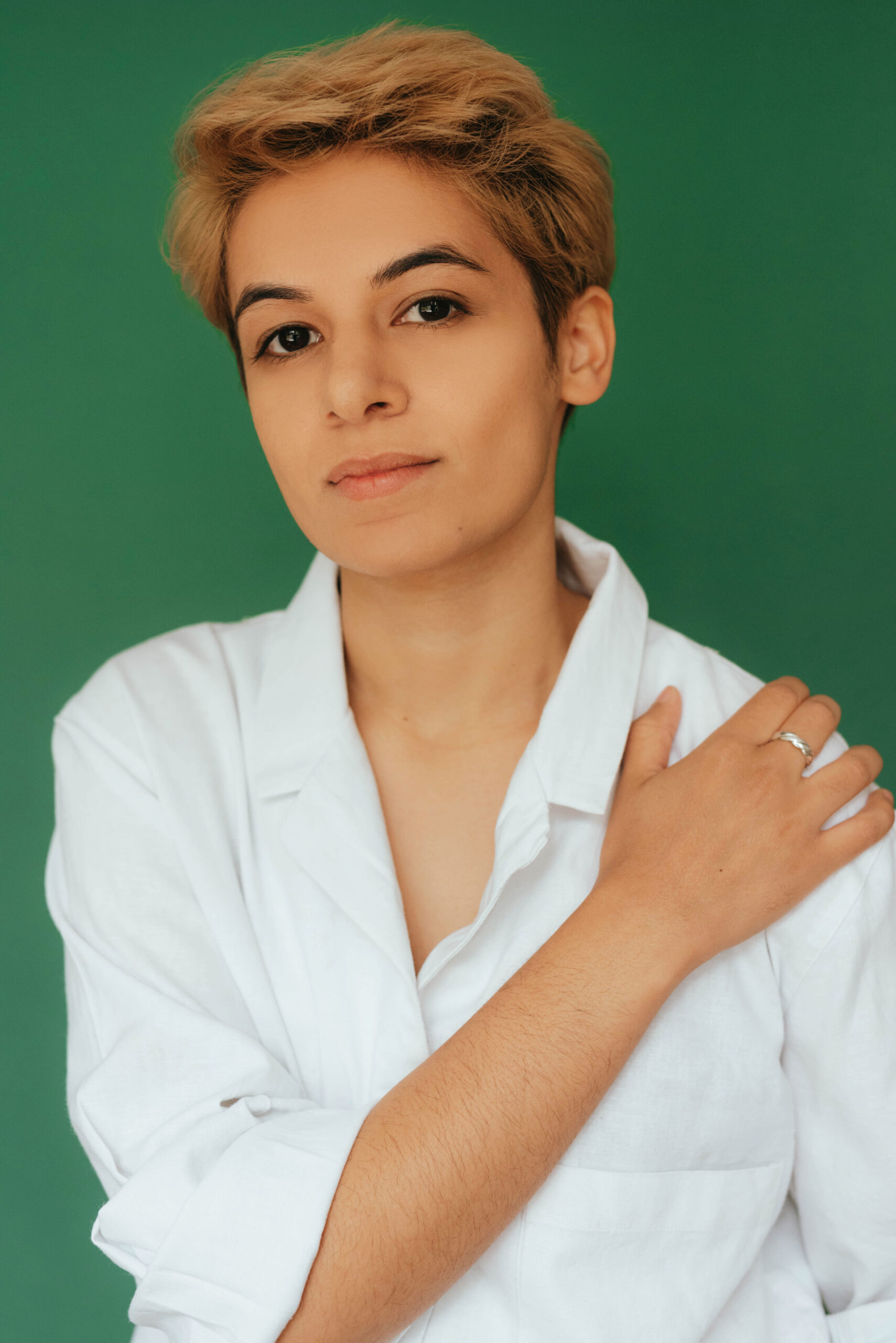Photo of Emerging Creators Unit Director Erum Khan - they have short dyed blond hair, and wear a white button down shirt. Their right hand rests on their left shoulder.
