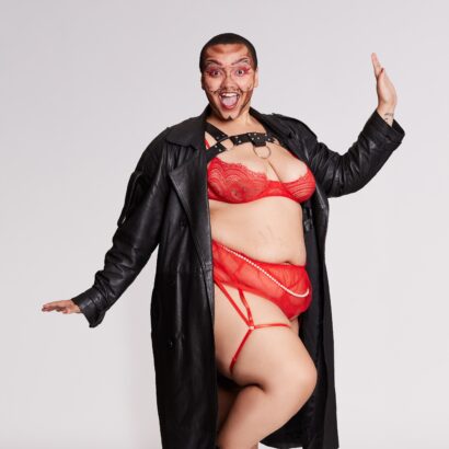 A fabulous photo of Sebastian URMOM, a mixed black androgynous presenting human with a curly buzzcut. They are decked out in drag makeup, a sexy red lingerie set, and a black leather trench coat worn open.