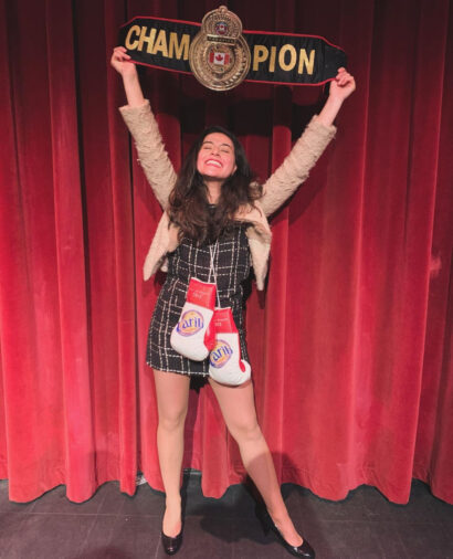 A photo of Dianna Aquilar on the Tallulah's stage. She is holding up a champion belt and boxing gloves hang around her neck.