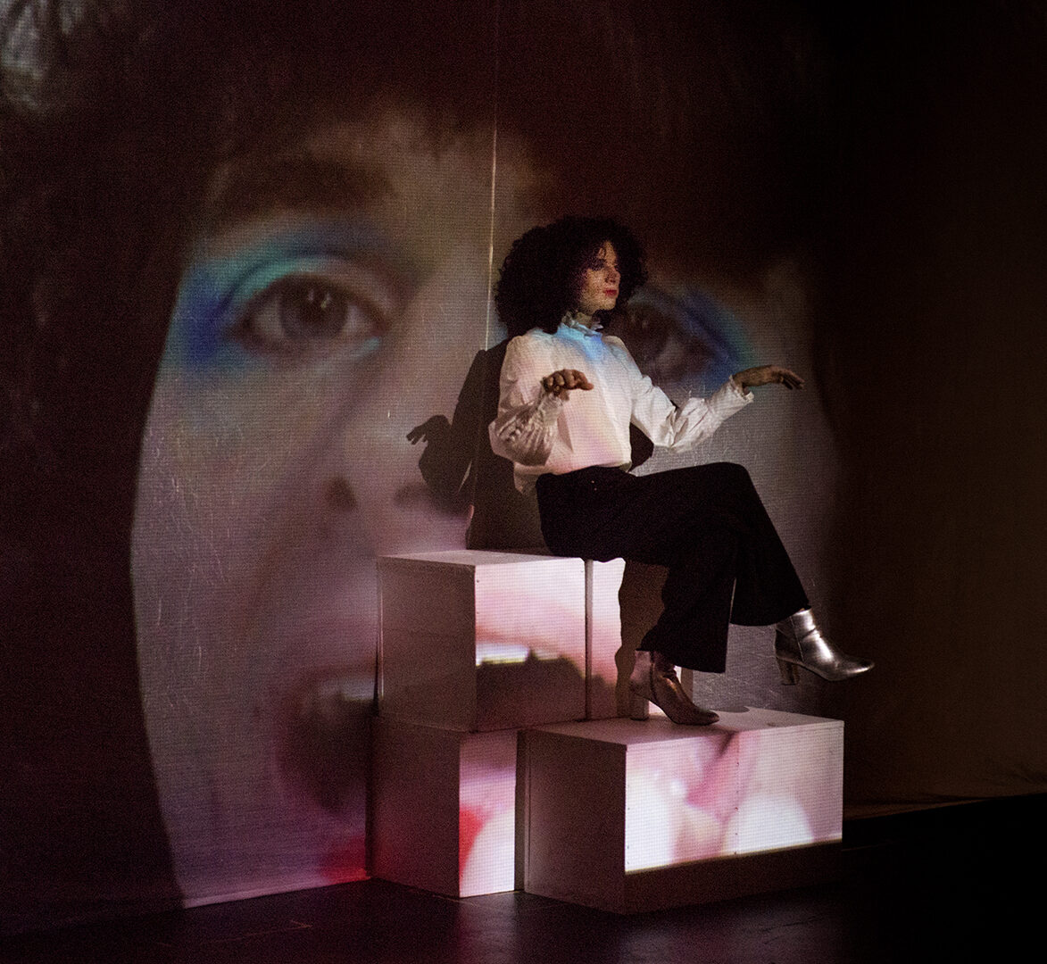 Image of James Knott on stage, sitting on white boxes, with a projected image of their face in the background. James is wearing a white long sleeve blouse, black pants, and shiny silver boots.