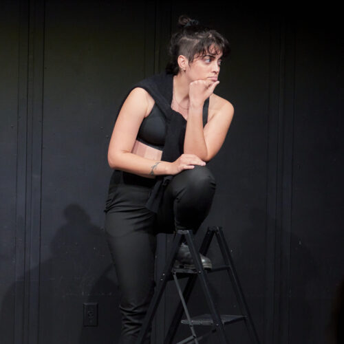 Ludmylla wears a black shirt and pants and stands with one leg up on a stepping stool.