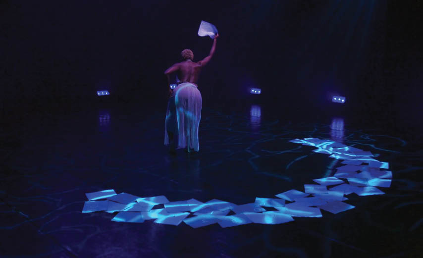 Image of Rhubarb artist Nickeshia Garrick on stage. Nickeshia is facing away, holding a piece of paper. Pieces of paper are scattered in a semi-circle along the edge of the stage space.
