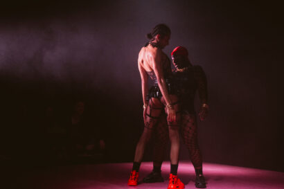 Two performers stand closely together on a dimly lit stage, creating an intense, intimate, and confrontational pose. Both are wearing bold, black fishnet bodysuits and edgy accessories, including gold jewelry. One performer wears a red cap and mask, while the other has braided hair and bright red shoes. The dark, smoky atmosphere adds drama, with faint audience figures visible in the shadows.