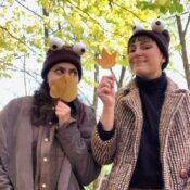 Image of two performers in hats with ping pong eyeballs on top. They are both holding fall leaves and wearing brown jackets.