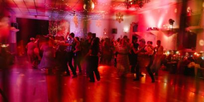 A red lit dancefloor of the owls club hall is filled with hot gay people dancing under a disco ball.