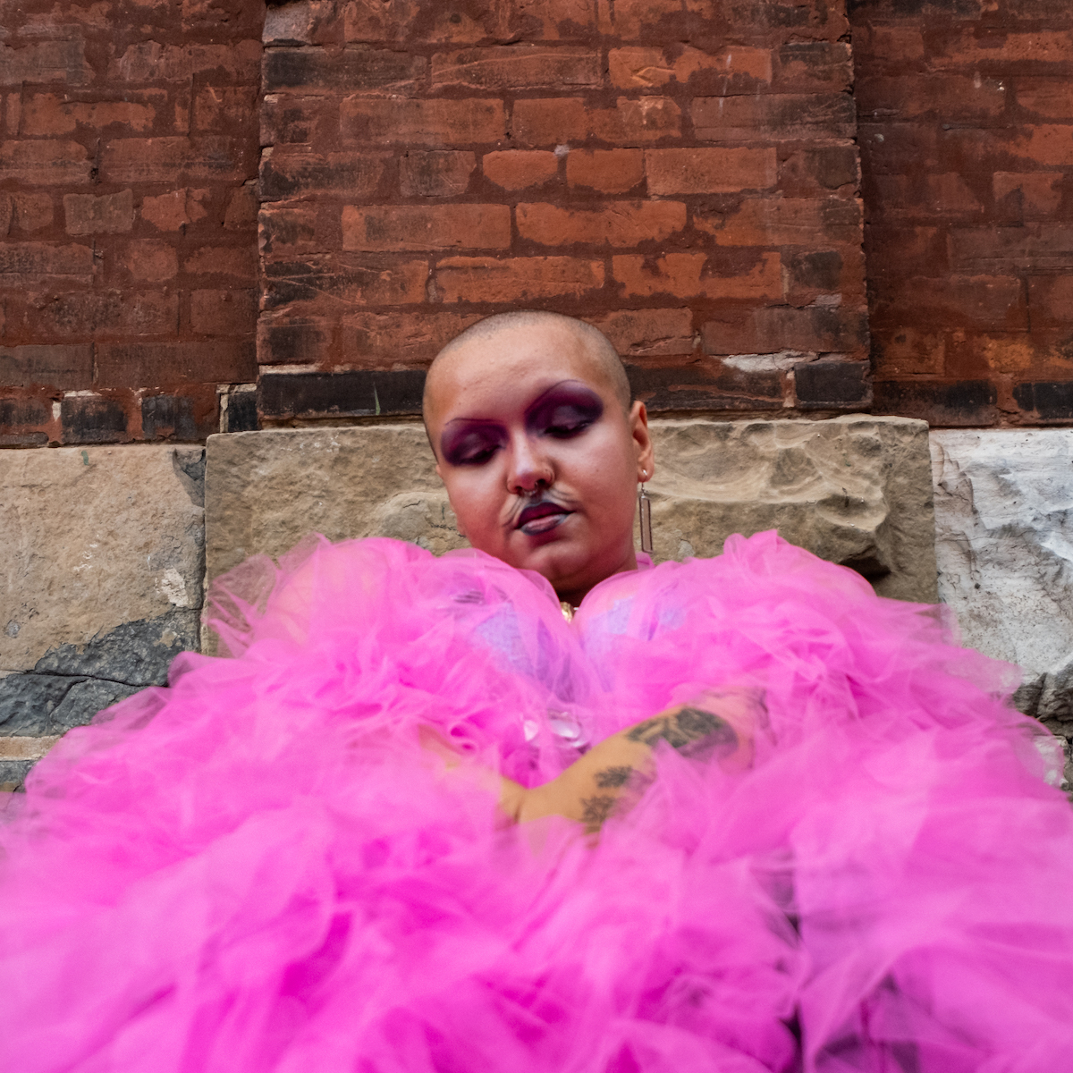 Photo of Sebastian Urmom - they have a shaved head and are wrapped in a pink crinoline. The background behind them is red bricks.
