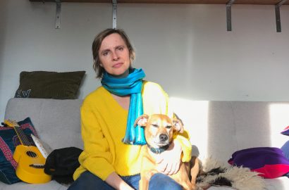 A woman (Evalyn's wife Suzie) wearing a yellow sweater and a blue scarf, sits on a couch with a brown dog (Fox).