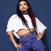 Image of Tygr Willy. They are wearing a white crop shirt and blue jeans, wearing drag makeup.
