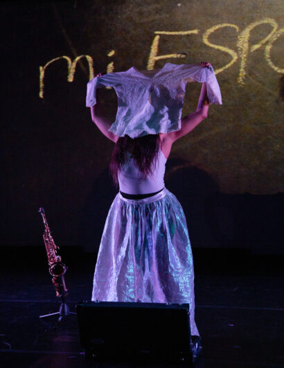 A performer stands on a dark stage, facing away from the audience, holding a white lace garment above their head with both hands. The figure wears a white tank top and a shimmering, translucent skirt that reflects stage lighting. A saxophone rests upright on the floor to the left. The backdrop displays the partially visible chalk-like text “mi ESP” in warm, earthy tones.