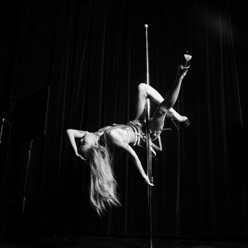 Image of Julie Phan, black and white photo of her dancing on a pole, wearing heels and a leather harness two piece. Julie has long hair.
