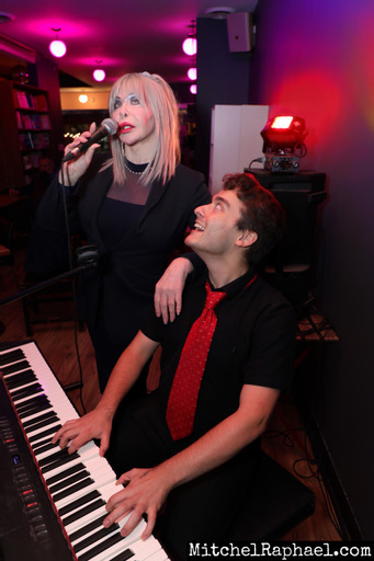 A light hearted photo of Mandy and Mateo in a room with pink lighting behind them. Mateo is a white man with short brown hair sitting at a keyboard and looking up at Mandy. Mandy is a White woman with shoulder length blonde hair singing into a handheld microphone. They are both dressed in all black with the exception of Mateo's bold red tie.