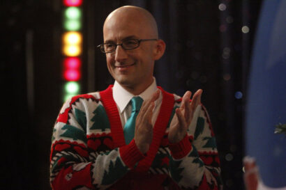 A festive photo of a white man with a bald head in front of some blurred christmas lights. He is wearing a red and green sweater and black plastic glasses.