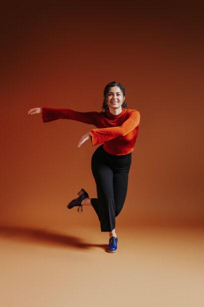 A tap dancer poses with her arms across her body to the side and her right leg in the air behind her left leg. She has long black curly hair in a ponytail and wears an orange top with flaired sleeves, black pants, and navy blue tap shoes. She poses in front of an orange gradient background -- as if she is about to break into tap dance.   